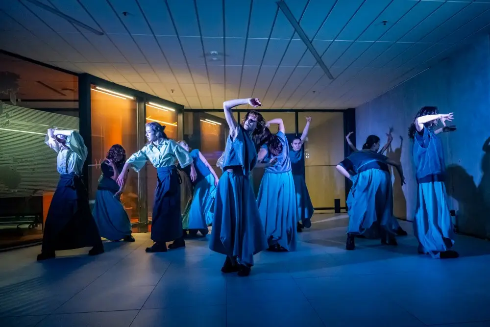 A group of dancers in loose blue costumes performs a synchronized routine indoors under dramatic blue and orange lighting, striking expressive, angular poses with raised arms.