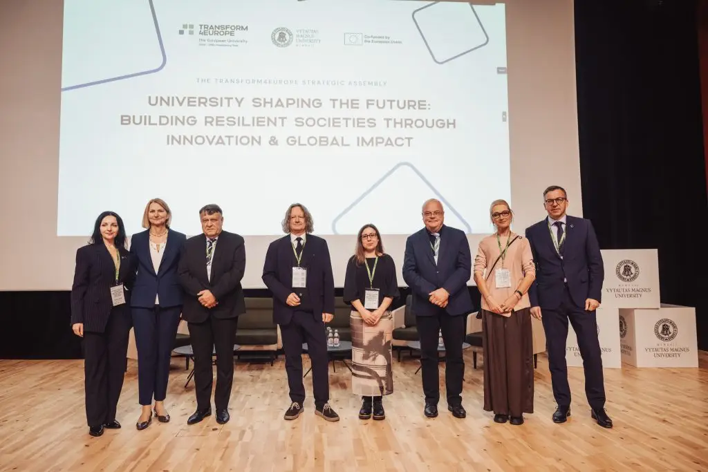 Eight professionally dressed people stand in a row on a stage, posing for a group photo in front of a large screen displaying the title of a university conference about innovation and global impact.