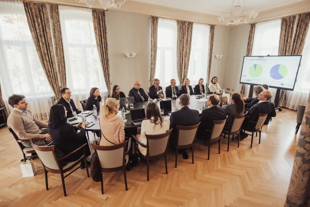 A group of people in business attire sit around a large conference table in a bright room, attentively watching a presentation with pie charts displayed on a screen.