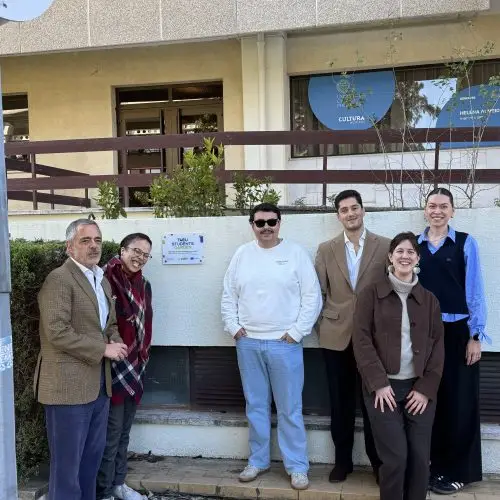 Six people stand and smile for a group photo outside a building with a plaque on the wall. The group is casually dressed and appears relaxed. There are plants and pavement in the foreground.