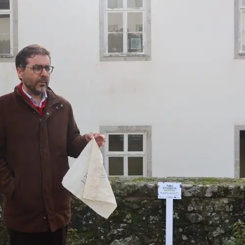 A man in glasses and a brown jacket stands outdoors holding a beige cloth, with a white building and a small sign on a post in the background.