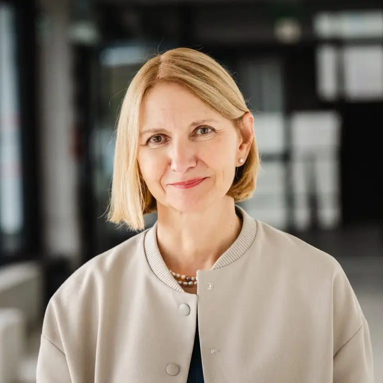 A middle-aged woman with blonde hair in a bob cut, wearing a light beige jacket, a dark top, and a pearl necklace, smiles slightly while standing indoors, embodying the spirit to Transform for Europe against a blurred background.