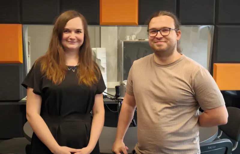 Two people stand side by side in an office with black and orange acoustic panels on the wall. One wears a black dress, the other a beige T-shirt. There are microphones and chairs in the background.