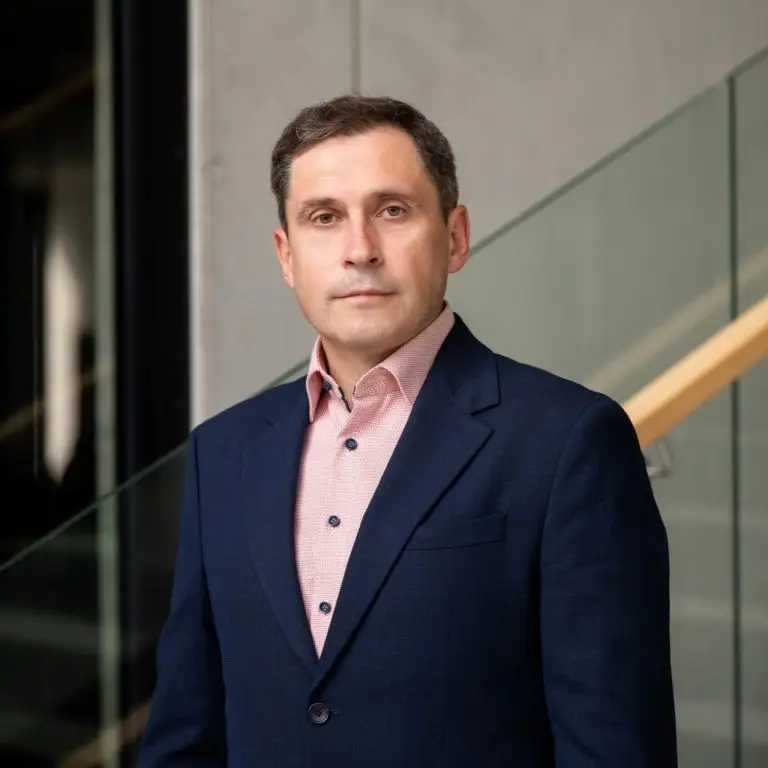 A man in a navy suit and light pink shirt stands indoors before a modern glass and wood staircase, looking directly at the camera with a neutral expression—poised to transform for Europe’s dynamic future.