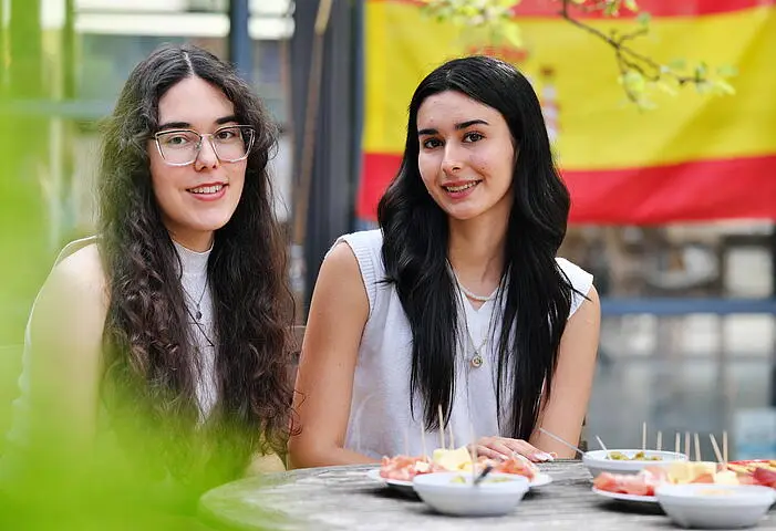Two young women sit at an outdoor table with plates of food in front of them. A Spanish flag hangs in the background. Both women are smiling and looking at the camera.