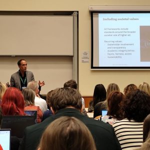 A man stands and speaks to a seated audience in a classroom. Behind him, a slide on a screen is titled Including societal values and lists topics like stakeholder involvement, equity, fairness, and sustainability.