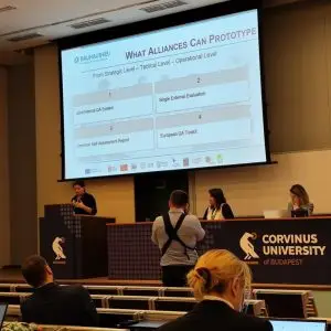 A speaker presents at a Corvinus University of Budapest event, with three people seated at a panel. A slide titled “What Alliances Can Prototype” is projected behind them. Audience members are seated, listening.