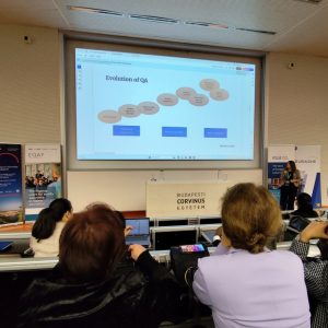 A speaker presents a slide titled Evolution of QA to an audience in a lecture room at Budapest Corvinus University. Attendees are seated and facing the screen, listening attentively. Banners stand by the walls.