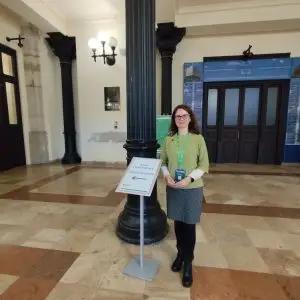 A woman with glasses, wearing a green top and gray skirt, stands indoors next to a sign that reads Data Humanities Registration, with a black column and ornate architecture in the background.