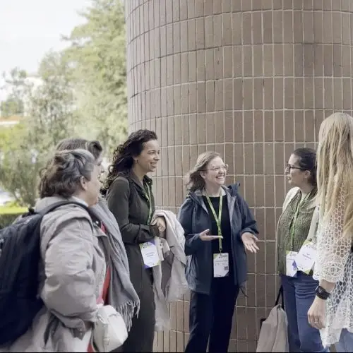 Five women wearing conference lanyards and name badges stand in a semi-circle outside, smiling and talking near a large cylindrical brick column. They appear to be enjoying a friendly conversation.