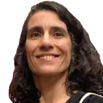 A woman with wavy dark hair, smiling, wearing a black top with a light-colored strap. The background is plain white.