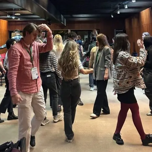 A group of people, some wearing conference badges, dance and interact in a modern room with wood-paneled walls and red chairs. A large screen displays information on one wall. The mood appears lively and social.