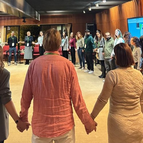 A group of people stands in a large circle indoors, holding hands. The room has wooden walls, a screen, and a few tables. Most participants are facing the center, engaged in a group activity or team-building exercise.