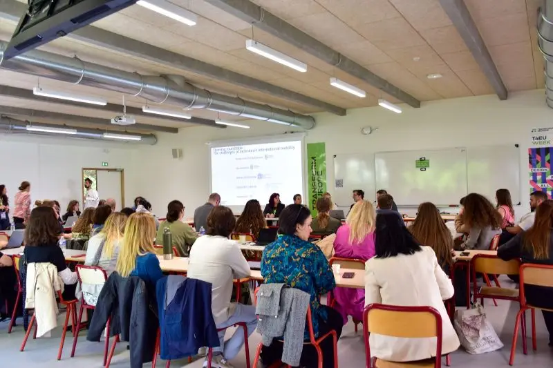 A group of people sit at tables in a classroom, facing a panel of speakers and a projected presentation. The room is brightly lit with modern decor and exposed ceiling pipes.