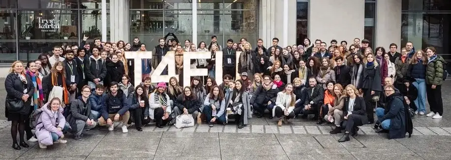 A large group of people pose outdoors in front of a modern building, standing and crouching around large white letters that spell T4F. Most are smiling and dressed in casual, winter clothing.