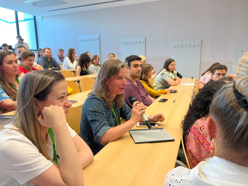 A woman in a classroom holds a microphone and speaks while others sit and listen attentively at long desks. The room is filled with people, some taking notes and others watching her.