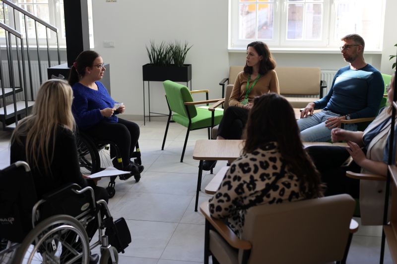 Five people, including two in wheelchairs, sit in a circle in a bright room having a discussion. Sunlight streams through large windows. The atmosphere appears collaborative and inclusive.