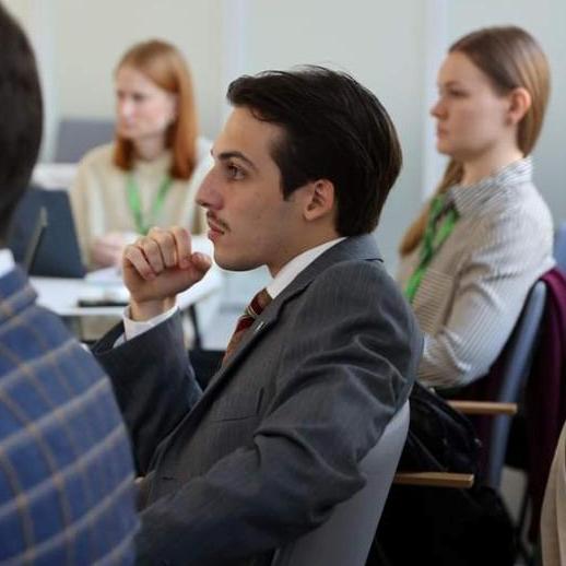 A young man in a suit sits attentively in a meeting room, resting his chin on his hand. Other people, also focused, are seated nearby with laptops and notepads visible.