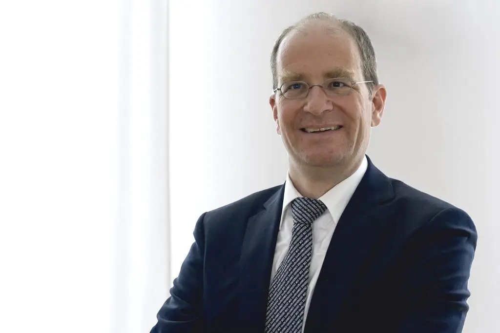A man in a suit, glasses, and patterned tie stands against a bright, white background, smiling with arms loosely crossed.