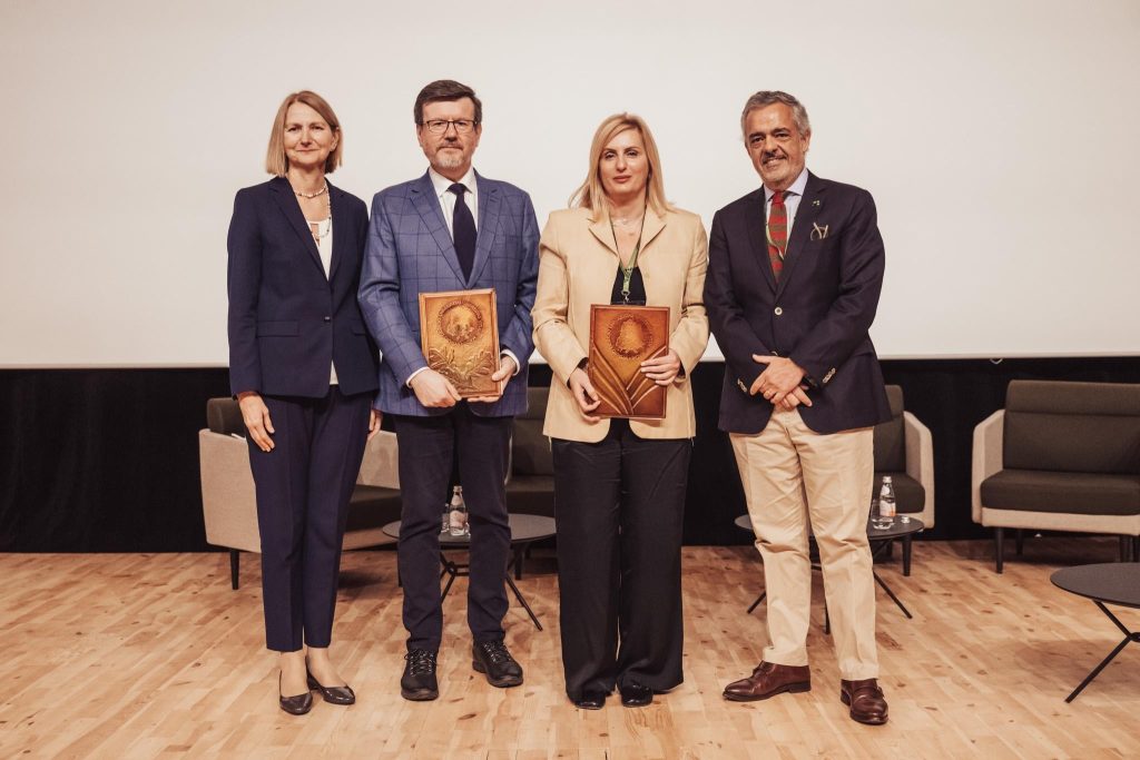 Four professionally dressed adults stand on a stage. Two people in the center hold plaques, while the other two stand on either side. There are chairs and a table in the background on a wooden floor.