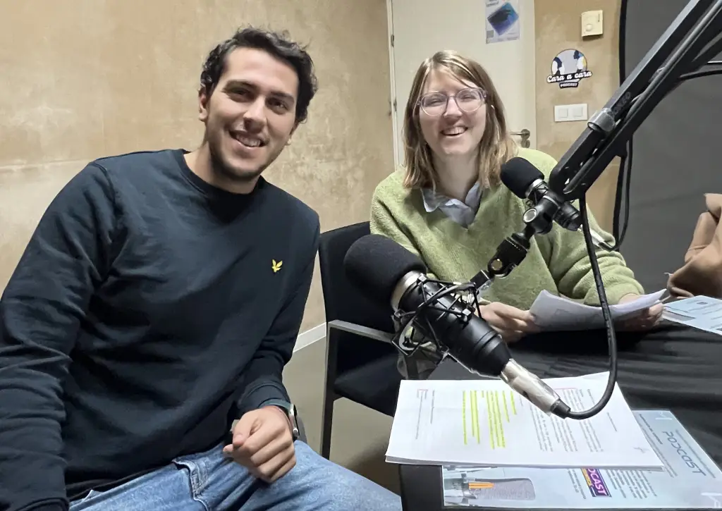 Two people sit at a table in a recording studio, smiling at the camera. They have papers in front of them and large microphones in front of each person, suggesting they are recording a podcast or radio show.