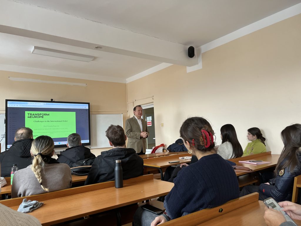 A man in a tan blazer stands at the front of a classroom speaking to students seated at desks. A presentation titled TRANSFORM EUROPE is projected on the screen behind him. Students listen and take notes.