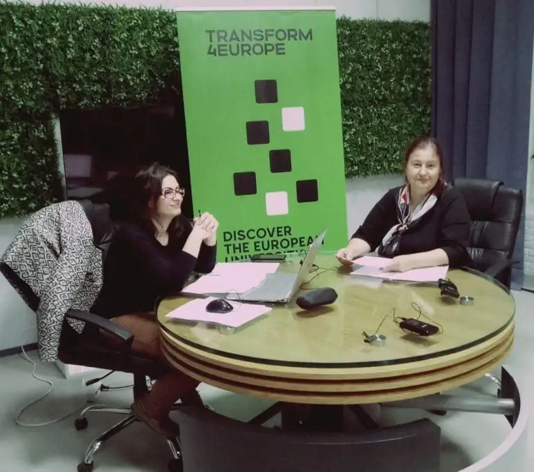 Two women sit at a round table with laptops and papers in front of a green Transform 4 Europe banner. The setting appears to be a modern office or meeting space with green wall panels.