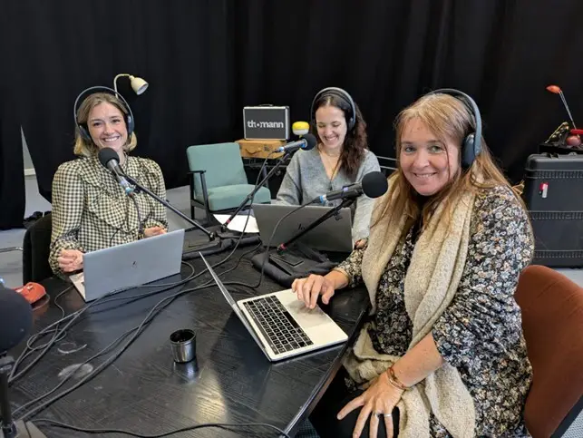 Three women with headphones sit at a black table with microphones and laptops, recording a podcast together. They are smiling and appear to be in a studio with a black curtain in the background.