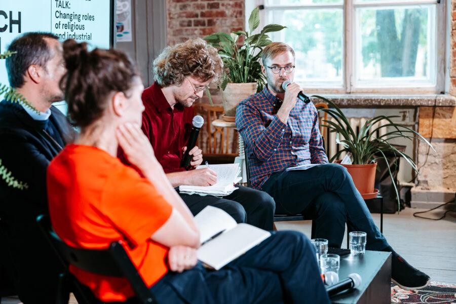 Four people sit on chairs indoors, engaged in a panel discussion. One man with glasses speaks into a microphone while others listen and take notes. Plants and large windows are in the background.