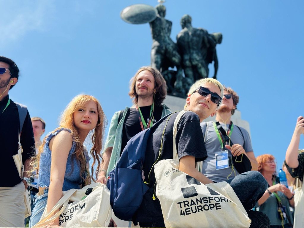 A group of young adults with tote bags that say TRANSFORM 4EUROPE pose and smile outdoors in front of a large statue under a clear blue sky.
