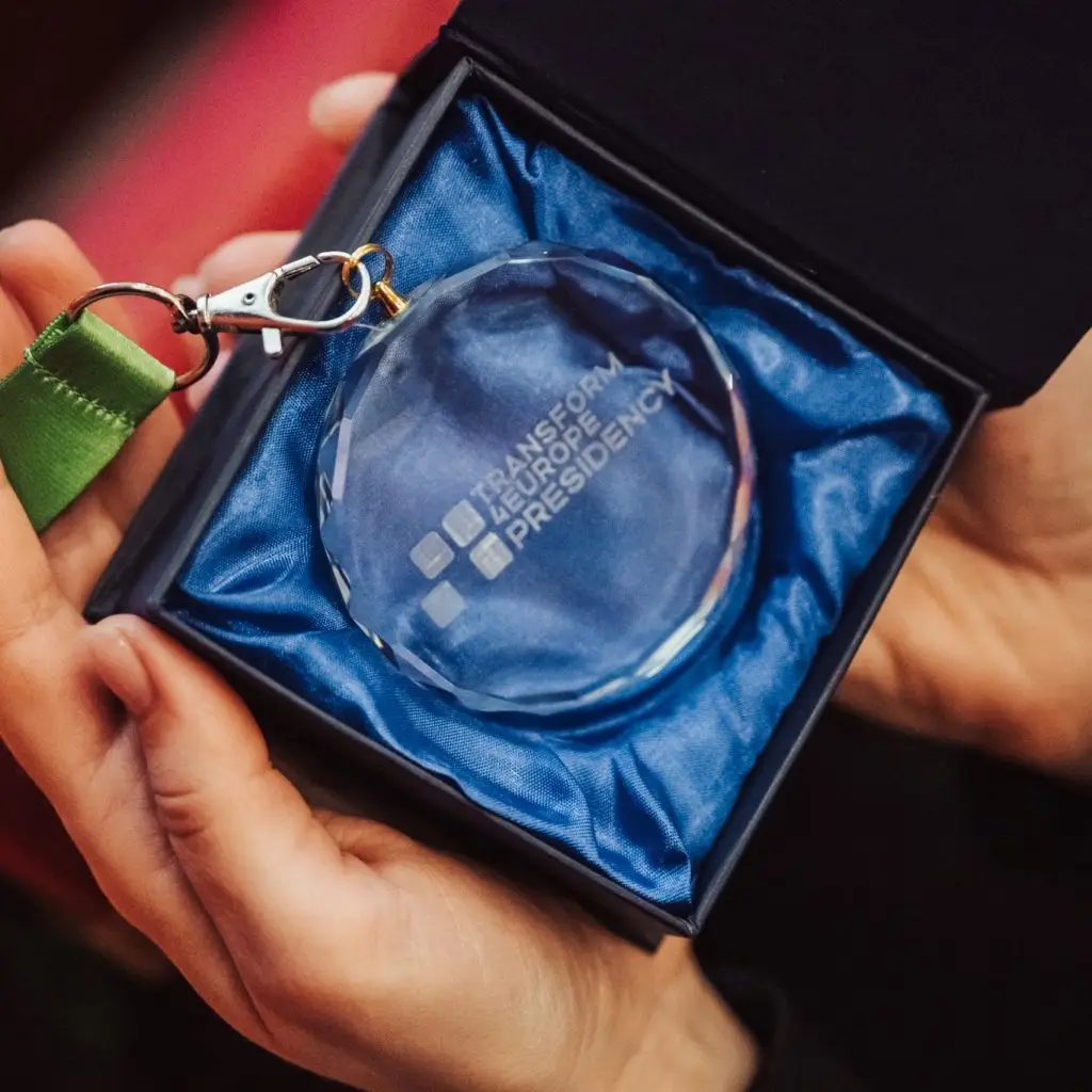 A person holds an open box containing a round glass keychain with the words TRANSFORM LEAD EUROPE PRESIDENCY engraved on it, resting on blue satin fabric.