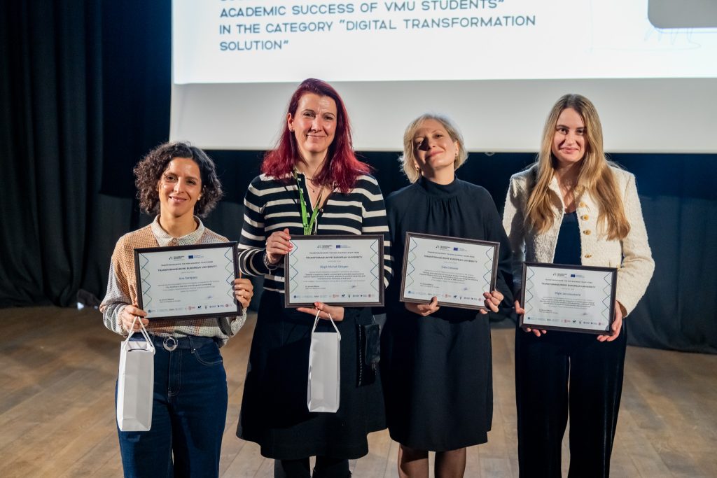 Four women stand side by side on a stage, each holding a certificate and a small white gift bag, smiling at the camera. A presentation screen is visible behind them.