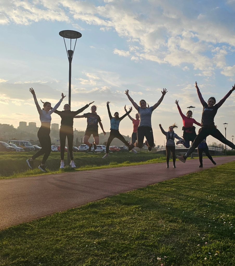 A group of people jump in the air with arms raised on a path in a park at sunset, with a city skyline, parked cars, and street lamps in the background. The sky is partly cloudy.