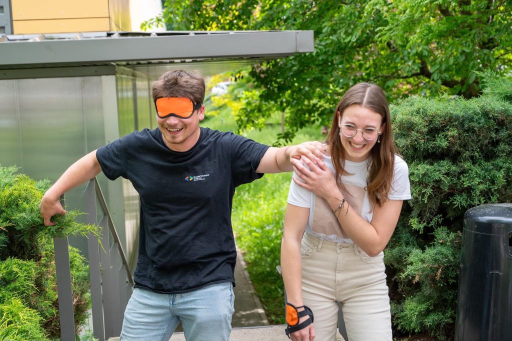 A smiling man wearing a black shirt and orange blindfold is guided up steps by a laughing woman with glasses and a white shirt, outdoors on a sunny day.
