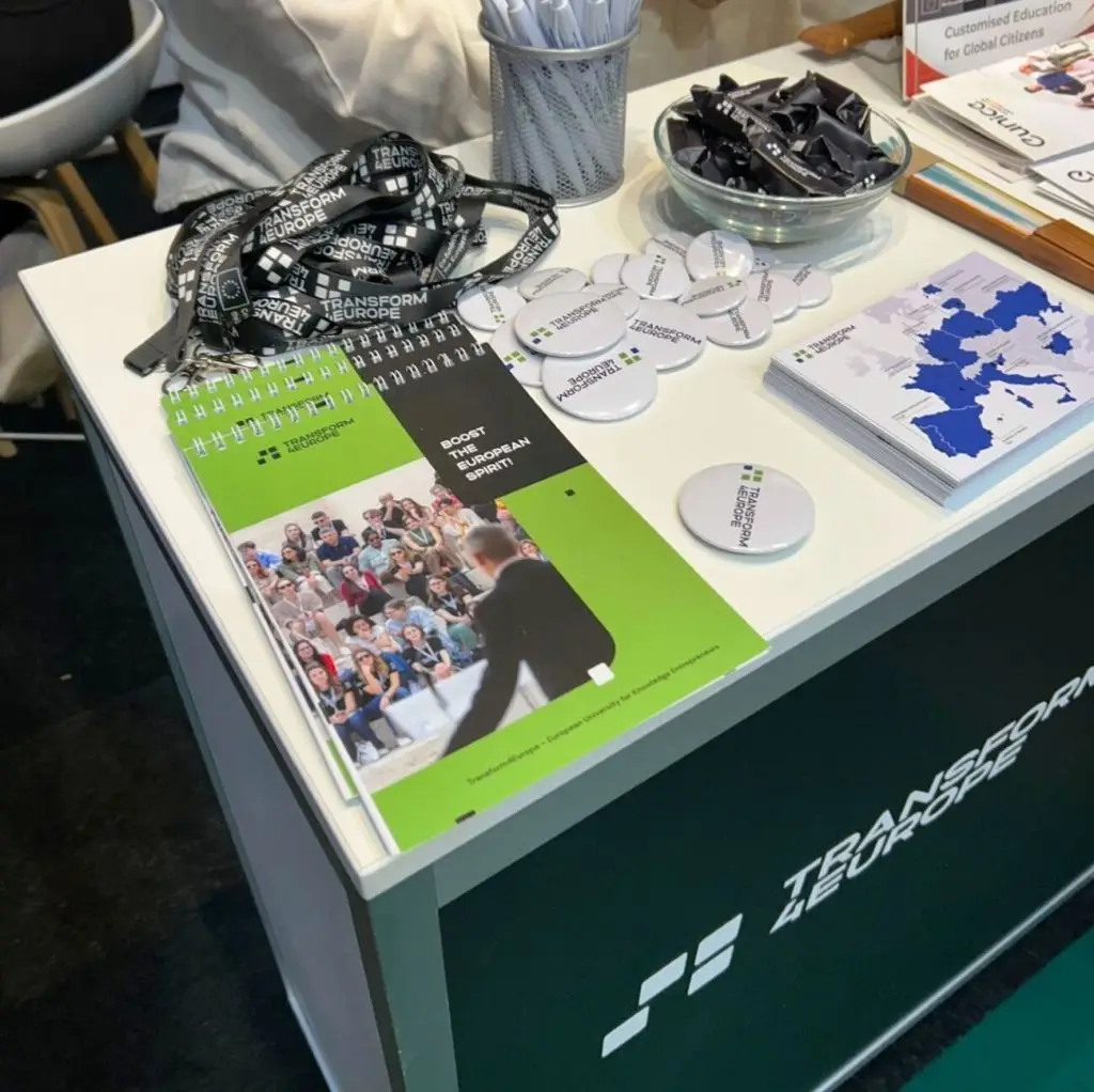 A display table with branded lanyards, buttons, pens, brochures, and a spiral-bound booklet. The table and materials are labeled Transform4Europe with a map and images of people in a conference setting.