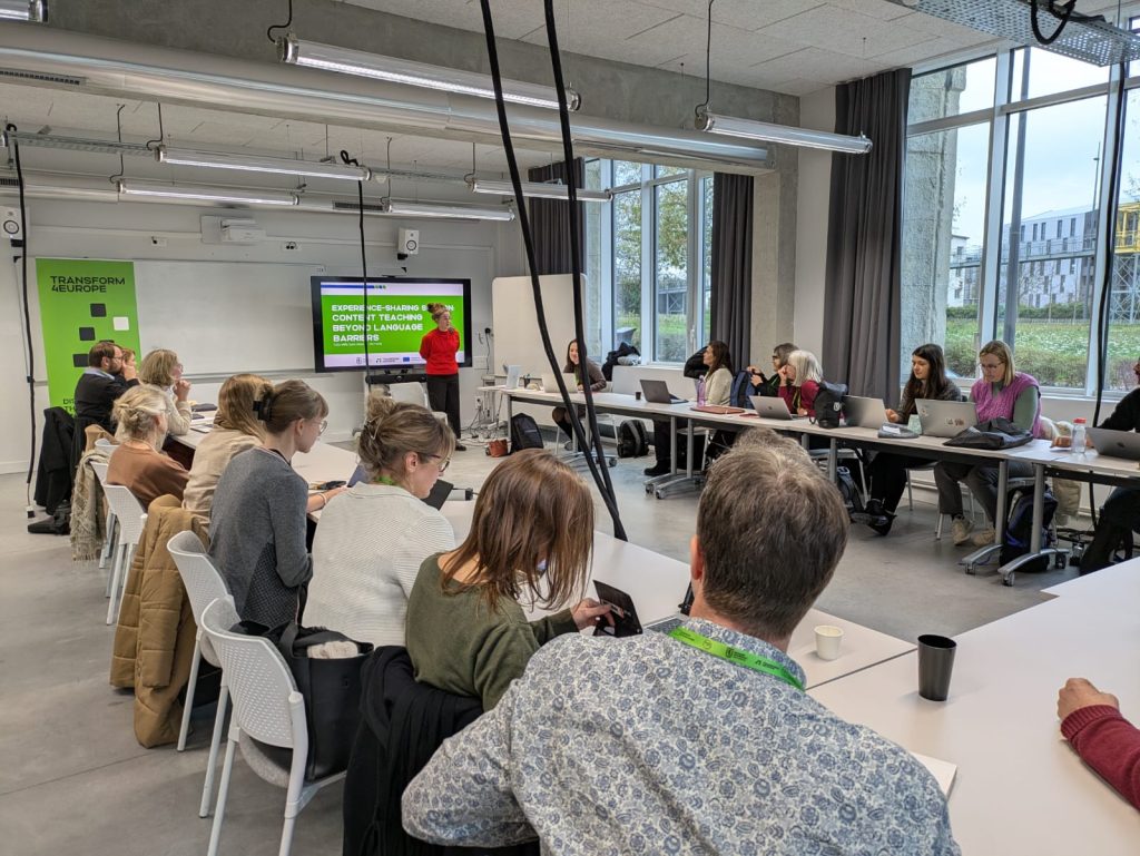 A group of people sit around tables in a bright conference room, listening to a presenter standing near a screen displaying a presentation. Laptops, notebooks, and drinks are on the tables. Large windows let in natural light.