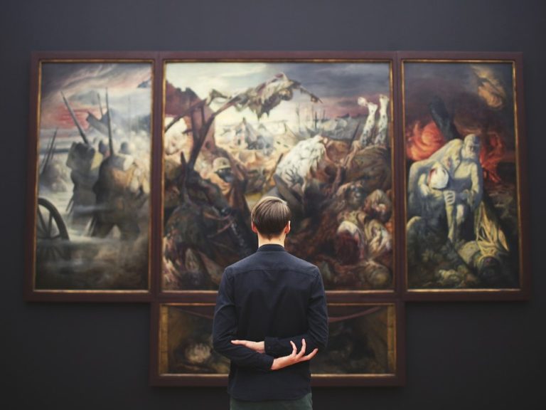 A person with short hair stands with arms behind their back, looking at a large, dark, three-panel painting depicting a chaotic battle or dramatic scene in an art gallery.