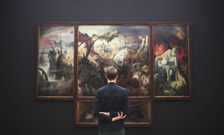 A person stands with arms crossed behind their back, viewing a large, dramatic triptych painting depicting a chaotic, historical battle scene in an art gallery.