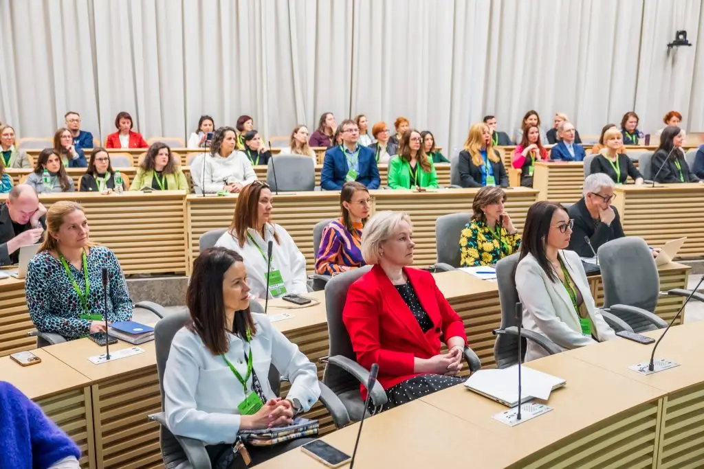 A large group of people, mostly women, sit attentively in a modern lecture hall with wooden desks and light-colored curtains, many wearing conference badges and green lanyards.