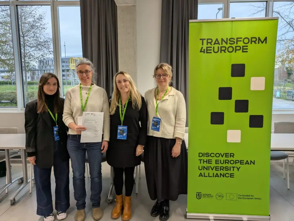 Four women stand indoors next to a green TRANSFORM4EUROPE banner. Three are smiling, and one holds a document. They wear badges and green lanyards, with tables and large windows in the background.