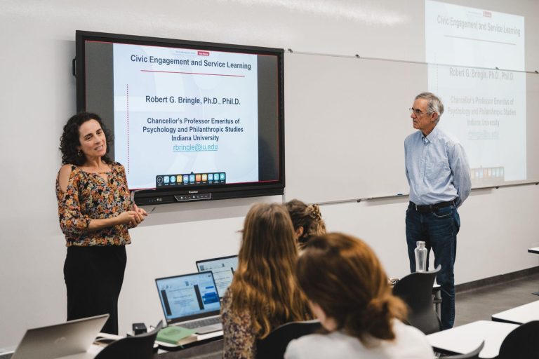 A woman stands and speaks to seated students in a classroom, while a man stands nearby. A presentation slide titled Civic Engagement and Service Learning is displayed on a large screen behind them.