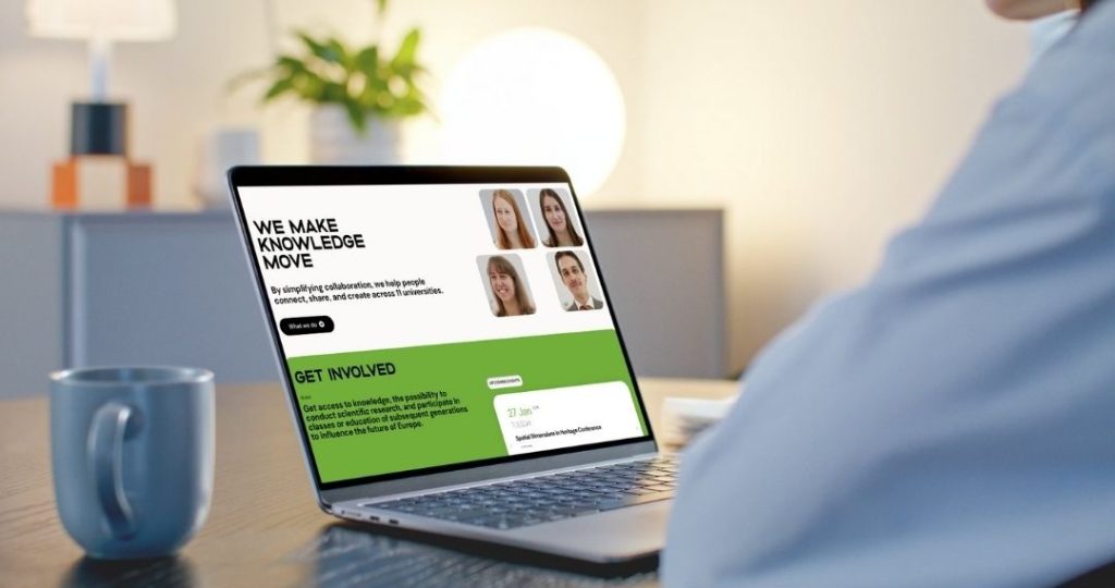 A person sits at a desk with a mug, viewing a laptop screen displaying a website titled We Make Knowledge Move with photos of four people and green sections for getting involved.