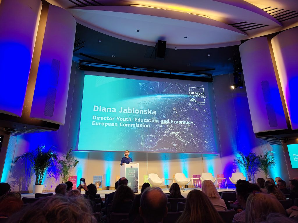 A woman stands at a podium on a stage with a large screen behind her displaying Diana Jablonska, Director Youth, Education and Erasmus+, European Commission. The audience is seated in a modern, blue-lit conference hall.