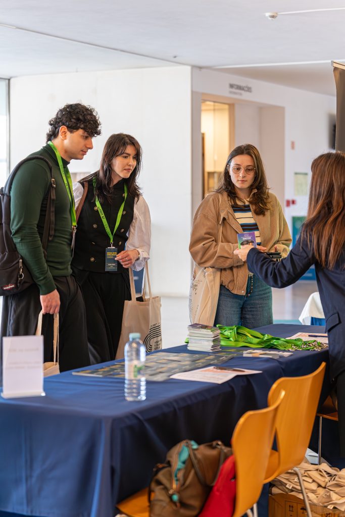 Three people stand at an information table covered with brochures, tote bags, and a water bottle, while a fourth person behind the table talks to them in a bright indoor setting.