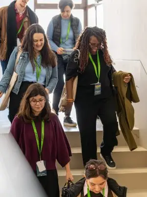A group of people wearing lanyards walk down a sunlit staircase, smiling and conversing. Some hold bags or jackets. The atmosphere appears casual and friendly.