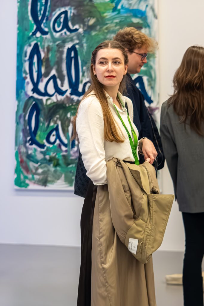 A woman with long brown hair, wearing a white blouse and holding a beige coat, stands indoors near abstract art with blue-green text. Two other people are in the background, partially turned away.