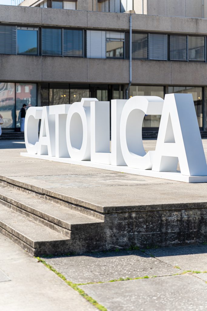 Large white letters spelling “CATÓLICA” are displayed outside a modern building with glass windows and concrete walls. Steps and pavement are visible in the foreground.