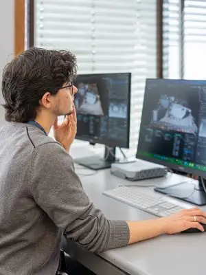 A person with dark hair and glasses sits at a desk, using a computer with two monitors displaying 3D modeling or graphic design software. They are looking thoughtfully at the screens.