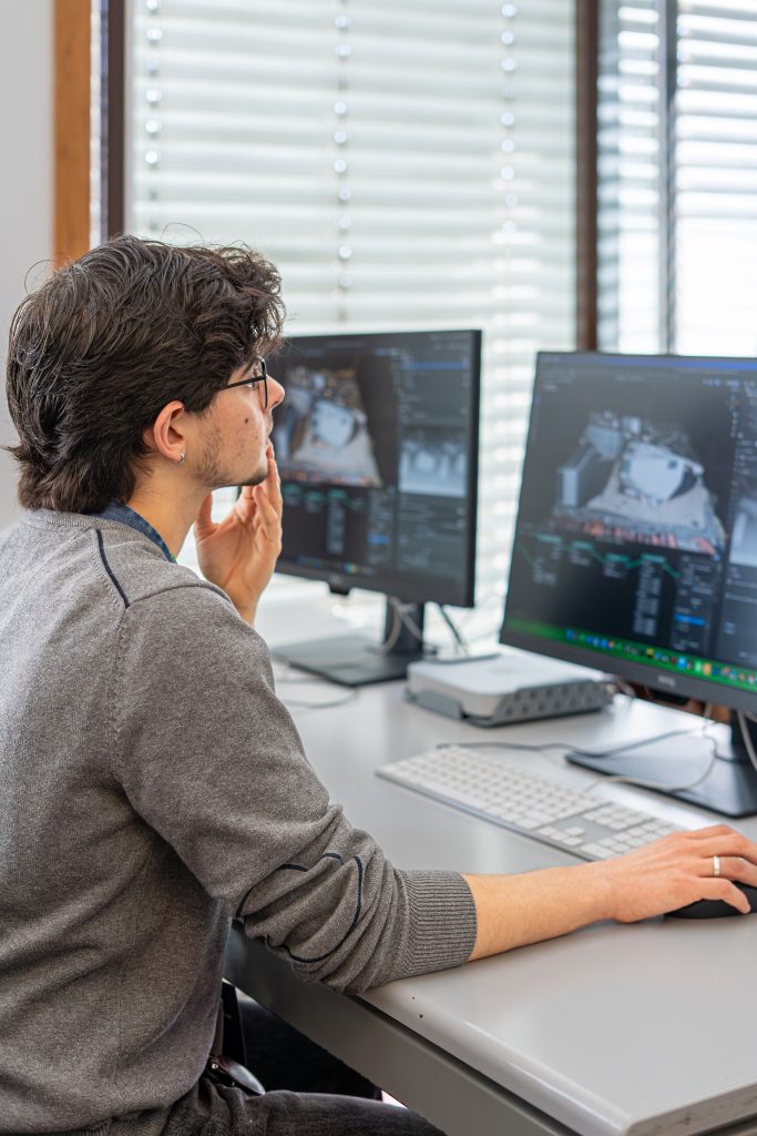 A person with dark hair and glasses sits at a desk using a computer with two monitors, analyzing or editing a 3D model or technical image, in a bright office setting with window blinds.