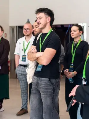 A group of people with green lanyards stand together indoors, listening attentively. The man in front wears a black t-shirt and gray jeans, smiling with arms crossed. Others stand behind him, also engaged.